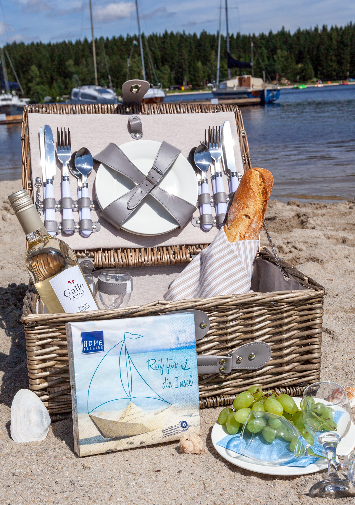 Ein Picknickkorb am Strand, prall gefüllt mit Geschirr, Servietten, Baguette, Trauben usw. verspicht einen schönen Nachmittag.