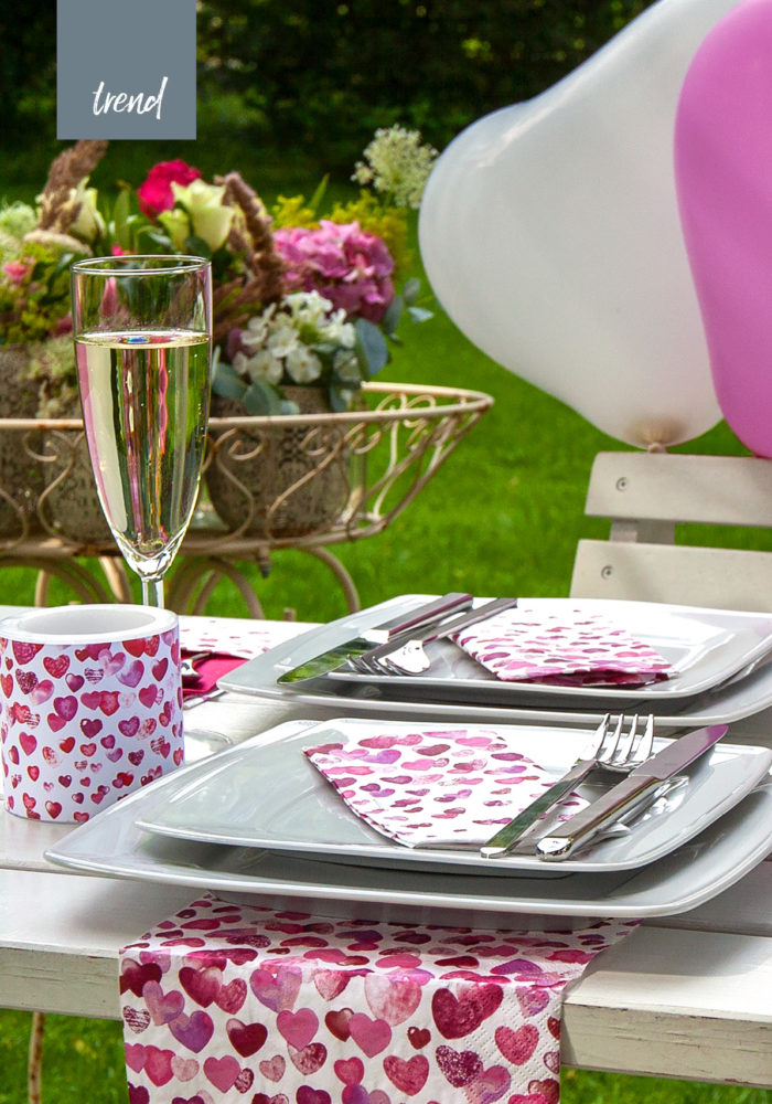 Ein bunt geschmückter Tisch mit Herzluftballons und passenden Servietten steht im Garten, alles ist hübsch dekoriert und die Gäste können kommen.