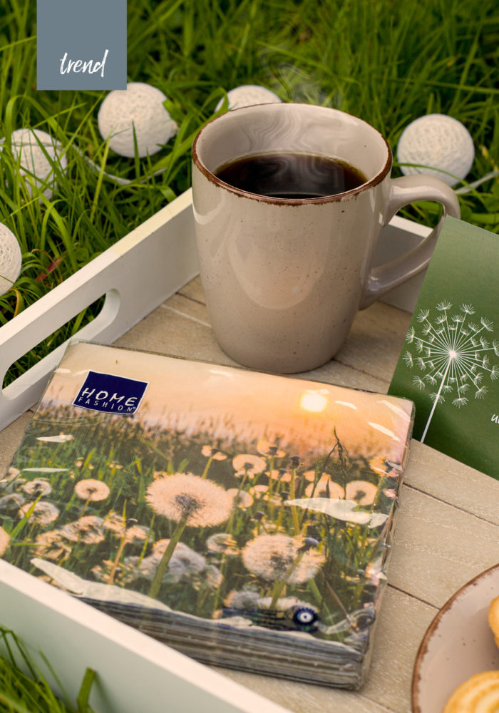 Ein Kaffee und Kuchen stehen auf einem Tablett bereit. Daneben eine Serviettenpackung mit Pusteblumen im Sonnenuntergang.