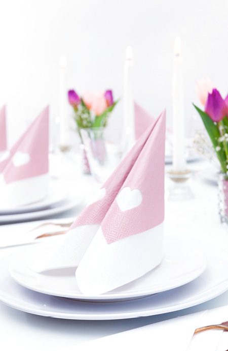 Eine rose farbige Serviette mit ausgestanztem Herz wird mit einer weißen Serviette im Tafelspitz gefalten und gibt der festlich gedeckten Tafel Schönheit und Eleganz.